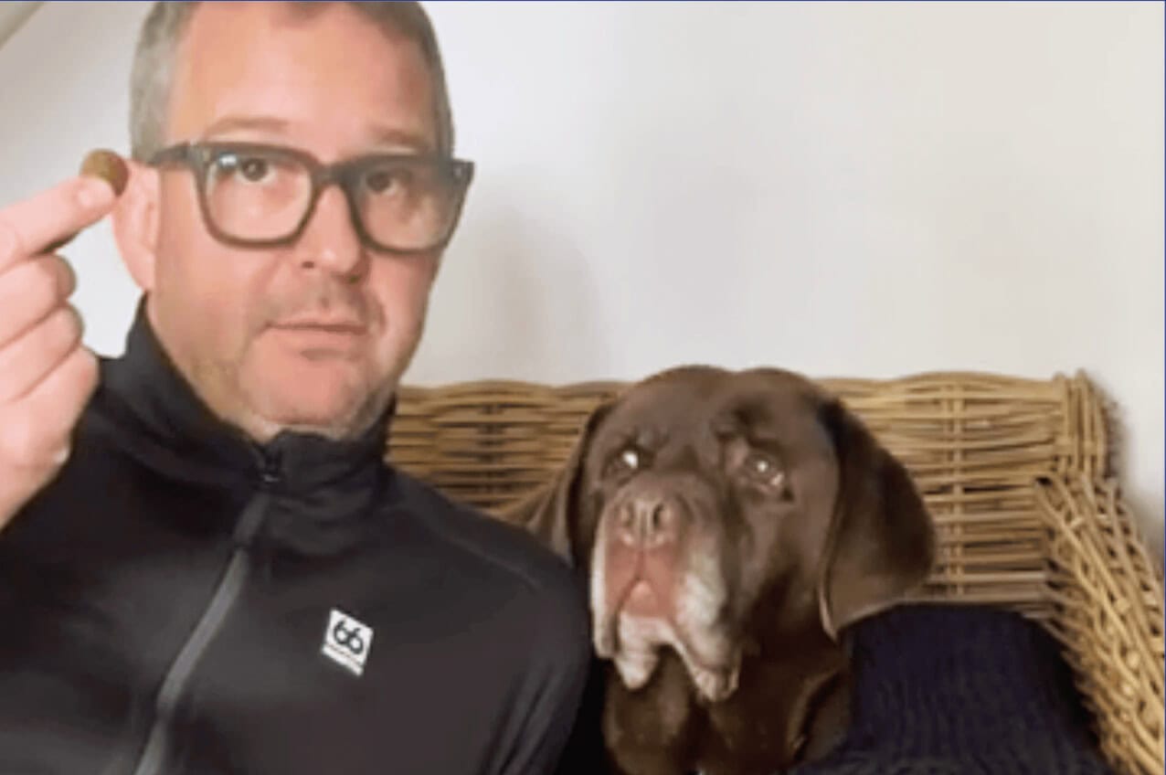 A man wearing glasses and a black jacket sitting next to a brown dog on a wicker sofa, both looking at the camera.