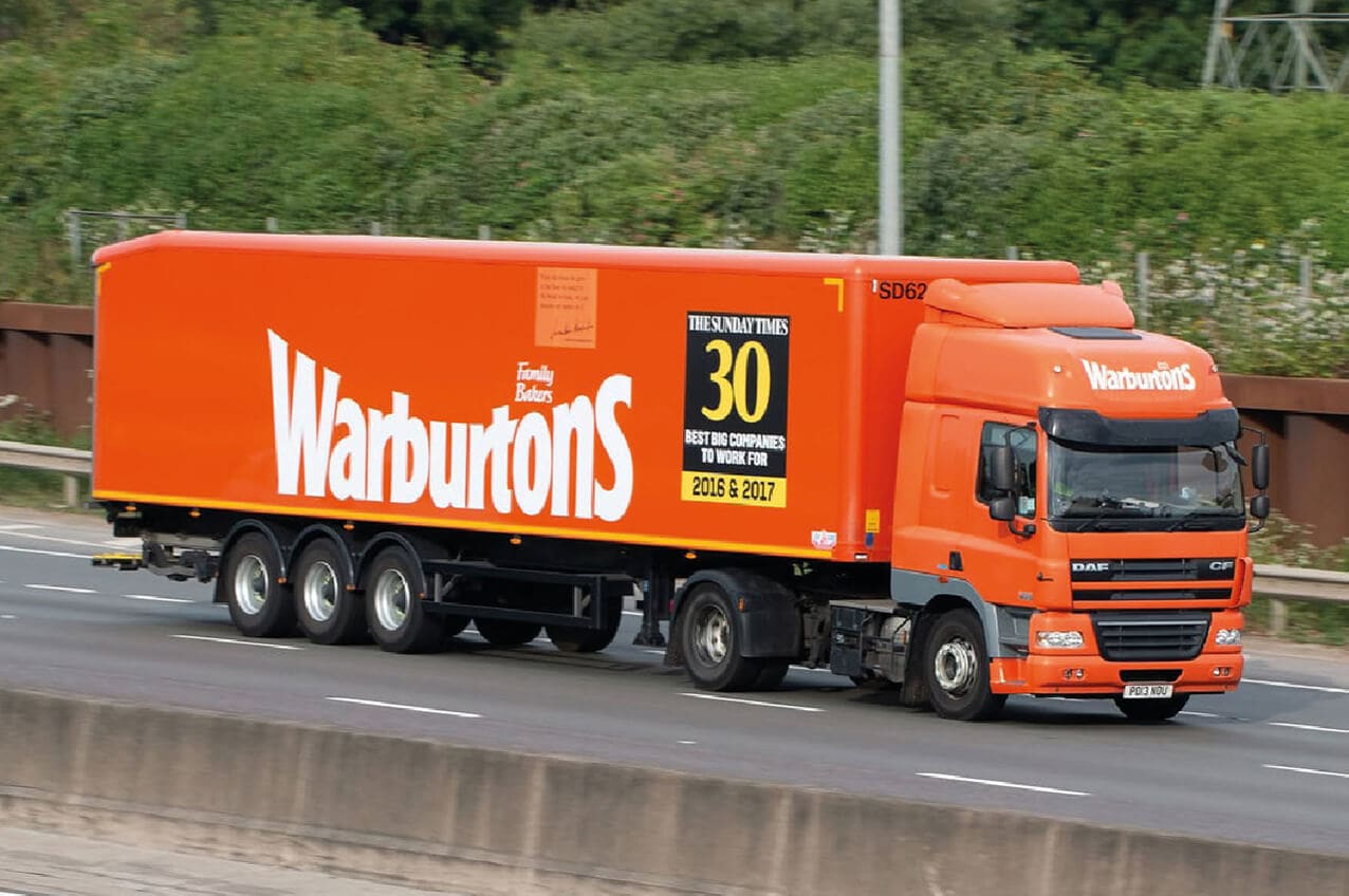 Bright orange Waburntons delivery truck on the highway with company branding and a promotional sign for 30 years of service.