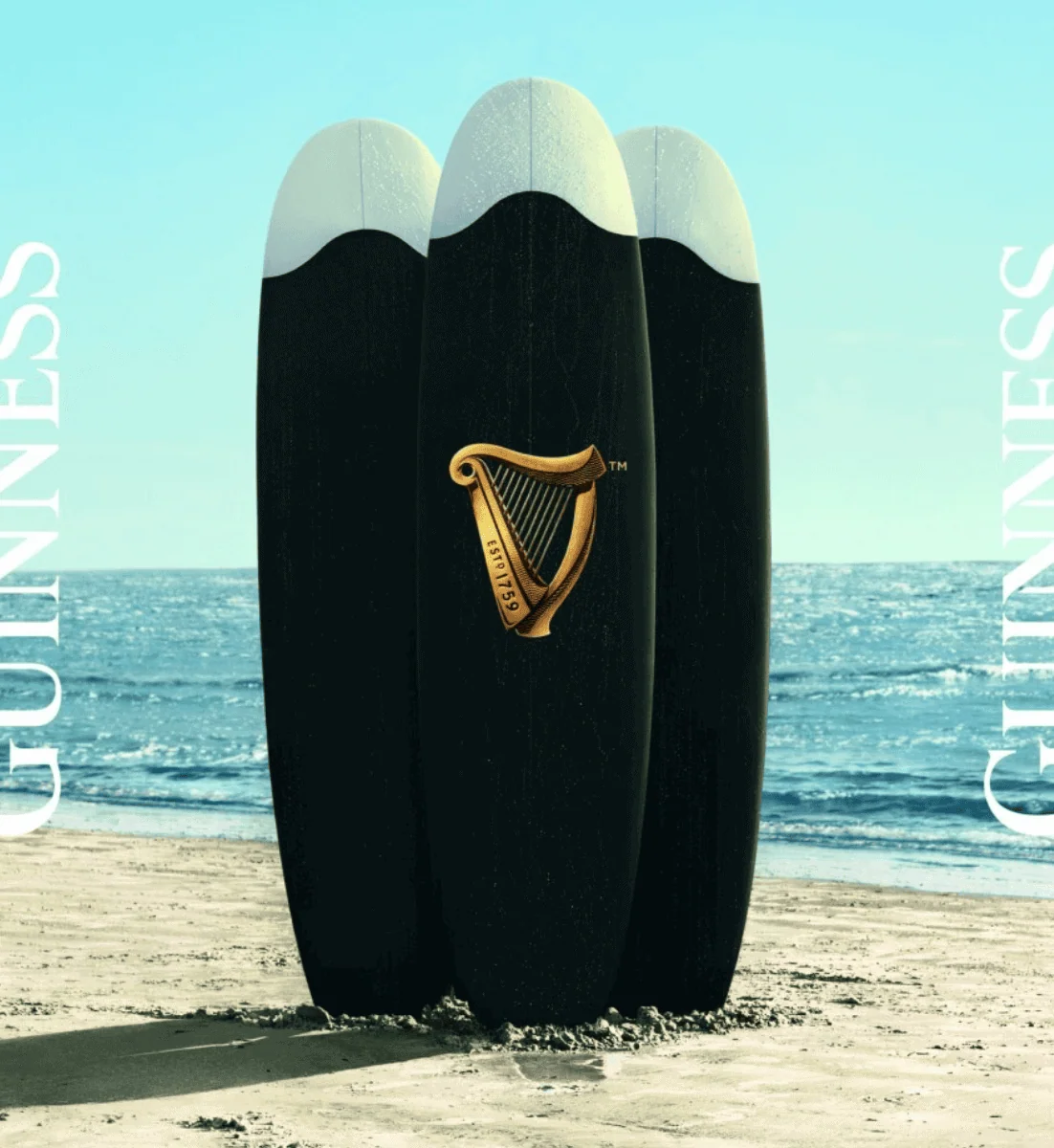 Surfboard shaped like a Guinness pint with harp logo, standing on sandy beach near the ocean, representing a creative branding concept for Guinness.