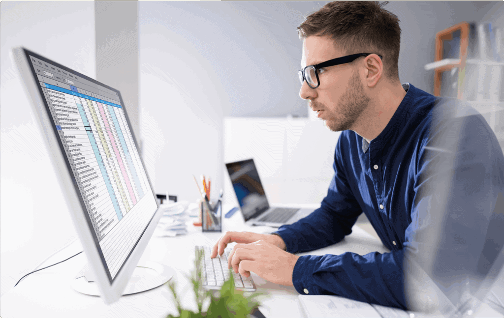 A young man with glasses analyzing financial data on a computer screen in an office setting.