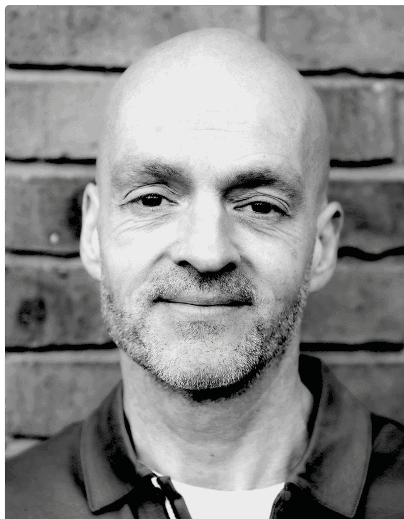 Black and white portrait of a smiling bald man with a beard, standing in front of a brick wall, representing professional financial leadership.