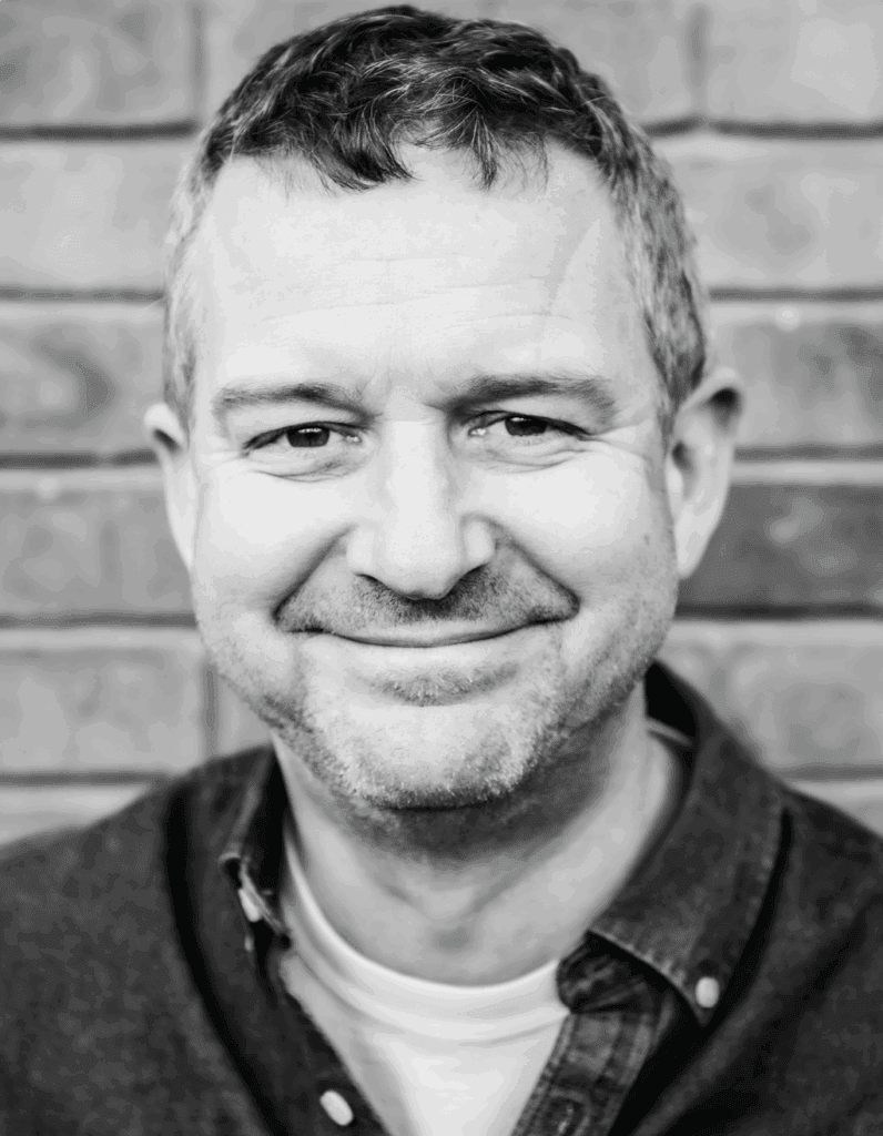 Close-up black and white portrait of a smiling man with short hair, wearing a casual shirt, standing against a brick wall background.